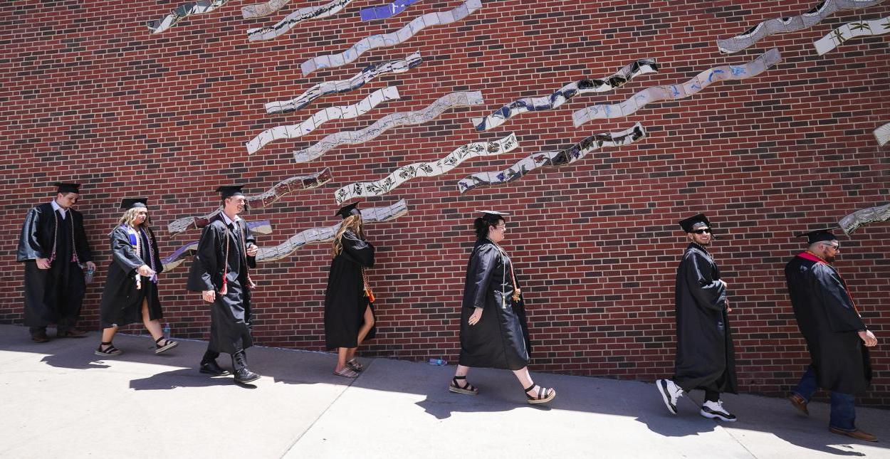 Seven students in black caps and gowns walk down a ramp in front of a brick wall showing a mural made of wavy tile.